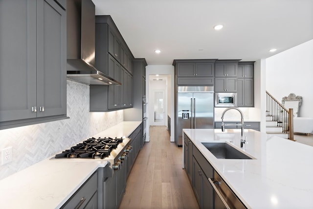 kitchen featuring light stone countertops, wall chimney range hood, sink, built in appliances, and light hardwood / wood-style floors