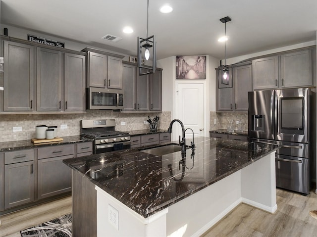 kitchen with appliances with stainless steel finishes, light wood-type flooring, tasteful backsplash, a kitchen island with sink, and pendant lighting