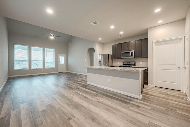 kitchen featuring ceiling fan, light stone countertops, an island with sink, appliances with stainless steel finishes, and light wood-type flooring