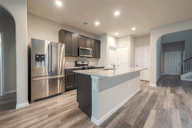 kitchen with appliances with stainless steel finishes, light stone counters, a kitchen island with sink, sink, and light hardwood / wood-style flooring