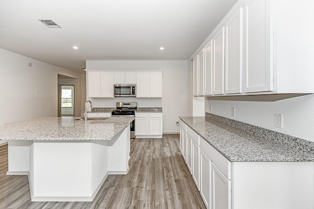 kitchen featuring stainless steel appliances, light stone counters, a center island with sink, white cabinets, and light wood-type flooring