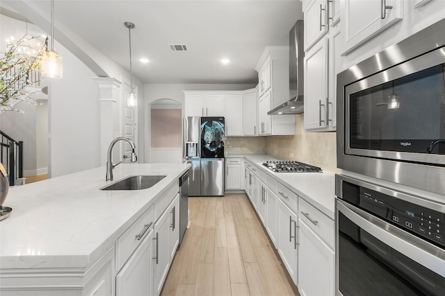 kitchen featuring arched walkways, light wood finished floors, stainless steel appliances, a sink, and wall chimney range hood