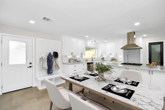 kitchen featuring white cabinetry, wall chimney range hood, tasteful backsplash, stainless steel dishwasher, and light tile patterned floors