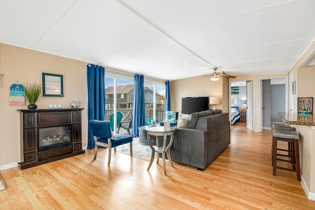 living room featuring ceiling fan, light wood-type flooring, a glass covered fireplace, and baseboards