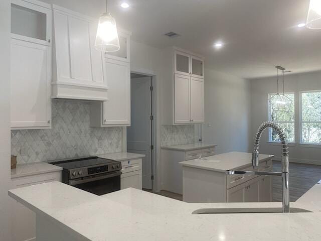 kitchen featuring stainless steel electric stove, sink, decorative light fixtures, white cabinets, and an island with sink