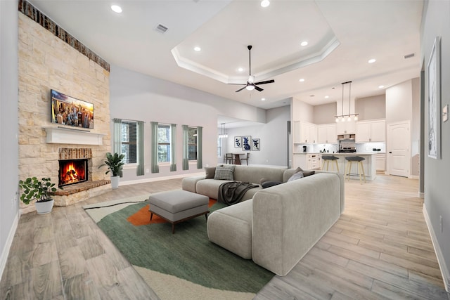 living room featuring ceiling fan, a stone fireplace, light wood-type flooring, and a tray ceiling