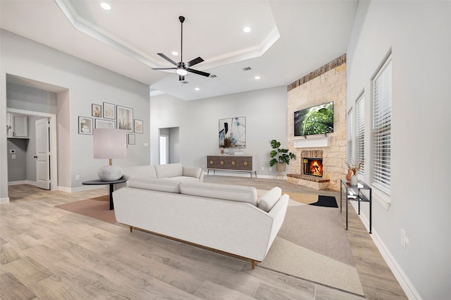 living room with a tray ceiling, a stone fireplace, ceiling fan, and light wood-type flooring