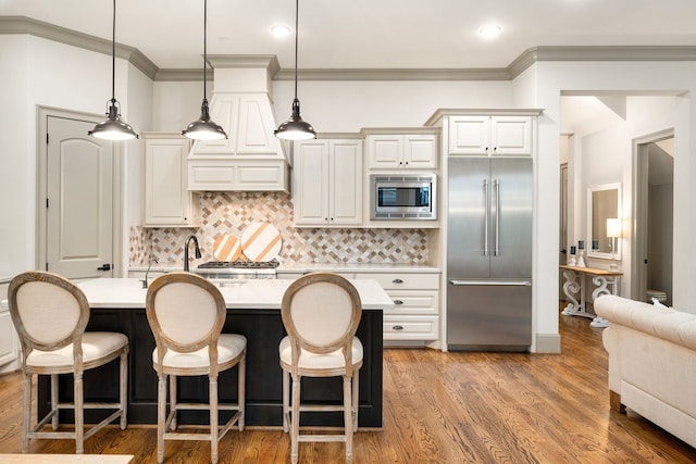 kitchen with tasteful backsplash, built in appliances, a kitchen island with sink, and hanging light fixtures