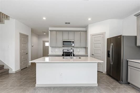 kitchen with appliances with stainless steel finishes, a kitchen island with sink, light countertops, gray cabinetry, and backsplash