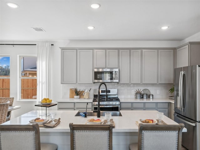 kitchen with gray cabinets, decorative backsplash, a center island with sink, and appliances with stainless steel finishes