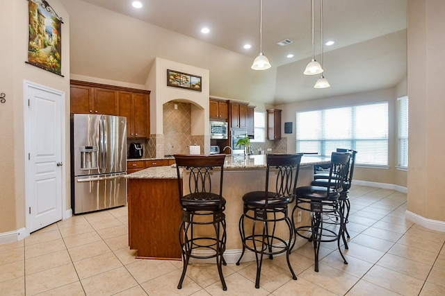 kitchen featuring a kitchen island with sink, stainless steel appliances, light stone counters, pendant lighting, and decorative backsplash