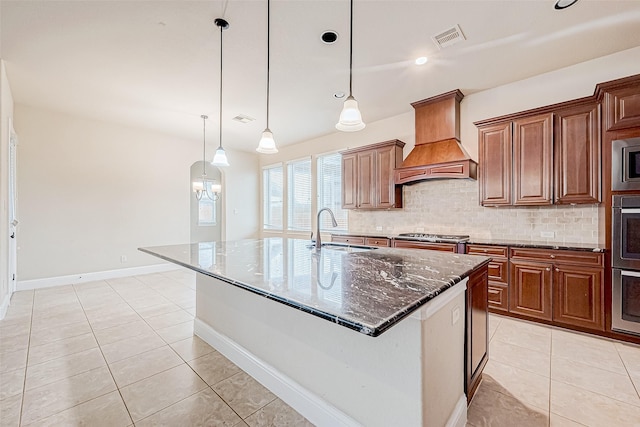 kitchen featuring dark stone counters, custom range hood, a kitchen island with sink, sink, and hanging light fixtures