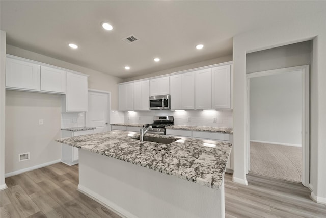 kitchen featuring appliances with stainless steel finishes, light wood-type flooring, backsplash, a center island with sink, and white cabinetry