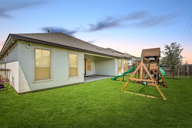 back house at dusk with a playground, a patio area, and a lawn