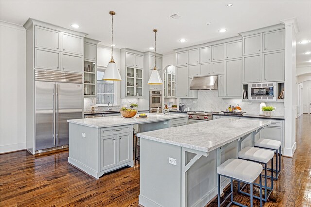 kitchen featuring dark hardwood / wood-style floors, built in appliances, pendant lighting, extractor fan, and a center island with sink