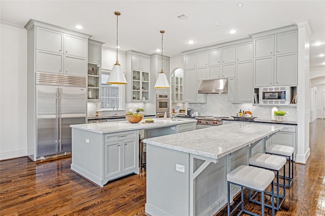 kitchen with an island with sink, built in appliances, light stone counters, and decorative light fixtures