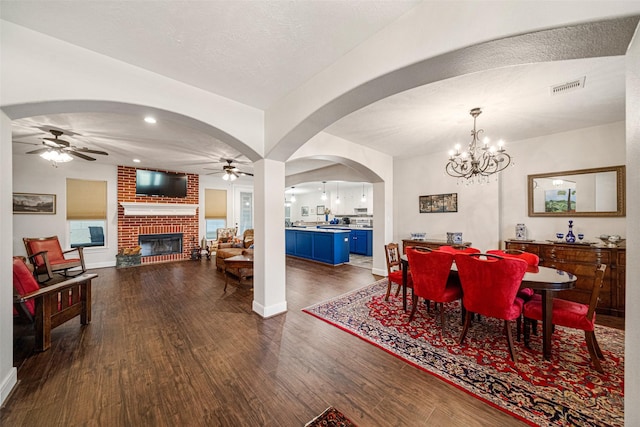 dining area featuring ceiling fan with notable chandelier, a textured ceiling, a brick fireplace, and dark wood-type flooring