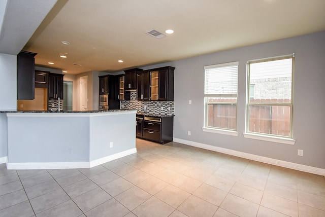 kitchen with stainless steel microwave, dark stone counters, decorative backsplash, light tile patterned floors, and kitchen peninsula