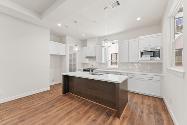 kitchen featuring decorative light fixtures, backsplash, stainless steel microwave, a kitchen island with sink, and white cabinets