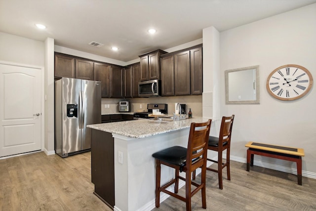 kitchen featuring stainless steel appliances, a kitchen breakfast bar, light stone counters, light hardwood / wood-style flooring, and dark brown cabinets