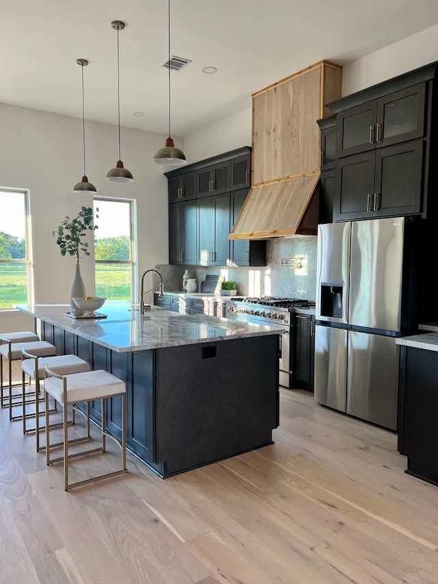 kitchen with appliances with stainless steel finishes, tasteful backsplash, a center island with sink, light hardwood / wood-style floors, and hanging light fixtures