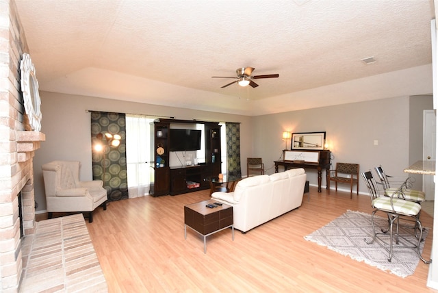 living room with a textured ceiling, light hardwood / wood-style floors, a brick fireplace, and ceiling fan