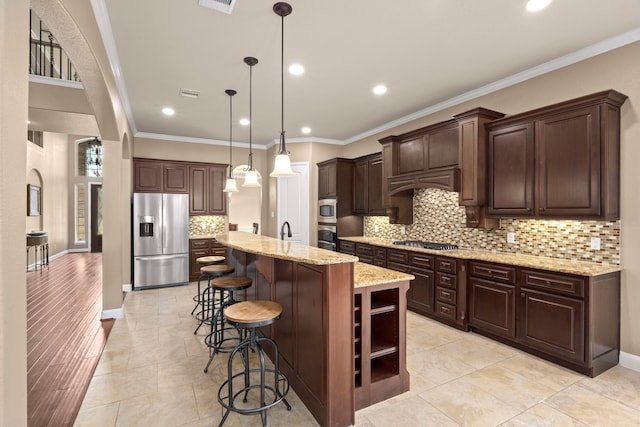 kitchen with pendant lighting, a kitchen island with sink, light stone countertops, ornamental molding, and stainless steel appliances