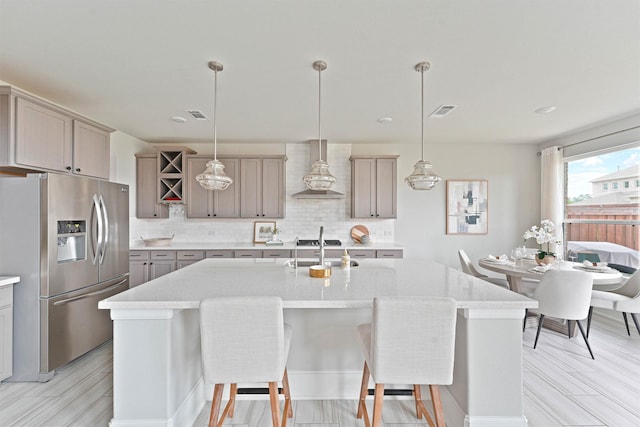 kitchen featuring stainless steel fridge, wall chimney exhaust hood, an island with sink, and decorative light fixtures