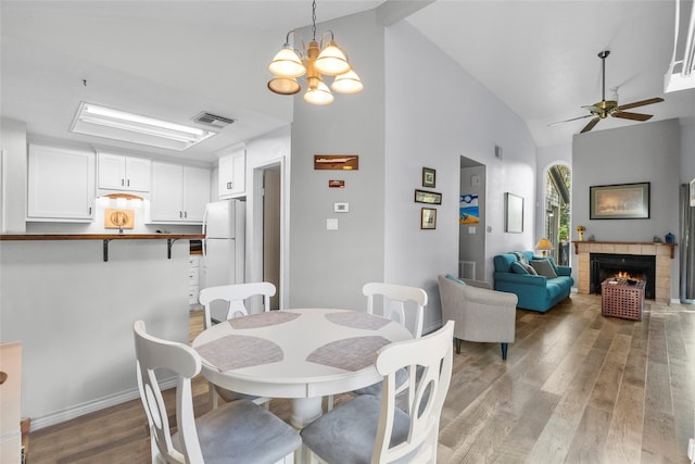 dining area with a tiled fireplace, light hardwood / wood-style flooring, high vaulted ceiling, and ceiling fan with notable chandelier