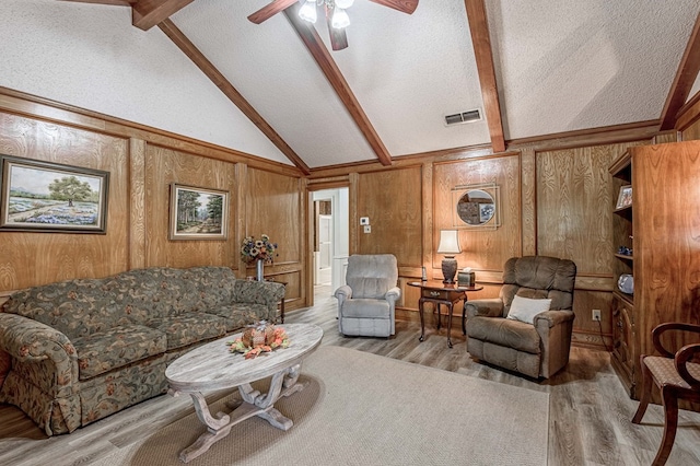 living room with light wood-type flooring, a textured ceiling, ceiling fan, wooden walls, and beamed ceiling