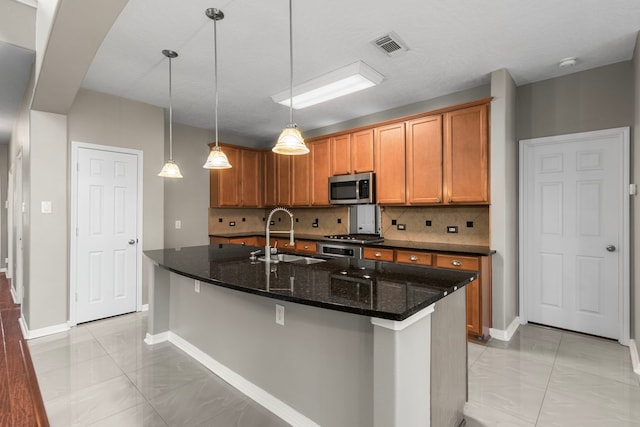 kitchen with tasteful backsplash, sink, dark stone countertops, hanging light fixtures, and an island with sink