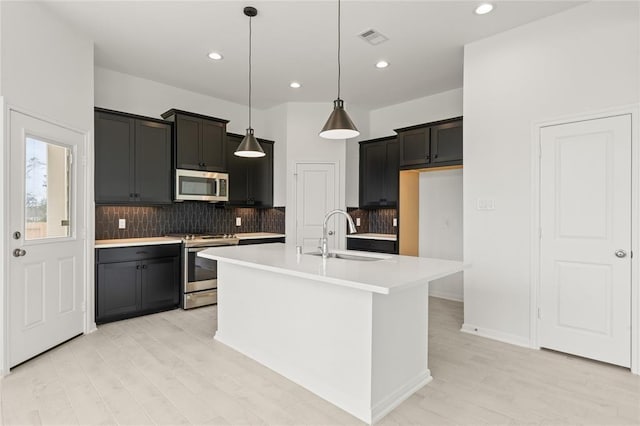 kitchen with sink, hanging light fixtures, stainless steel appliances, light hardwood / wood-style flooring, and a center island with sink