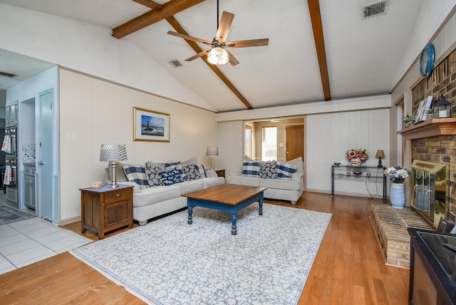 living room with a brick fireplace, a textured ceiling, ceiling fan, lofted ceiling with beams, and light hardwood / wood-style floors