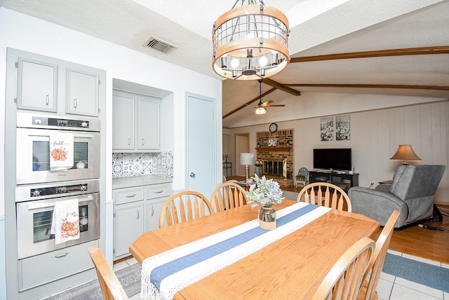 dining area featuring a brick fireplace, lofted ceiling with beams, light hardwood / wood-style floors, wooden walls, and ceiling fan with notable chandelier