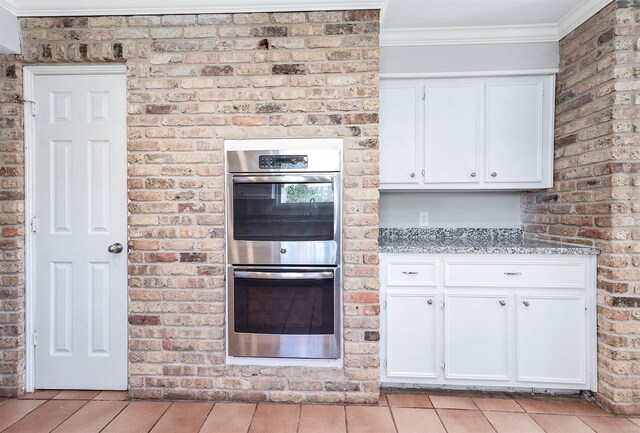 kitchen featuring light stone counters, brick wall, double oven, crown molding, and white cabinets