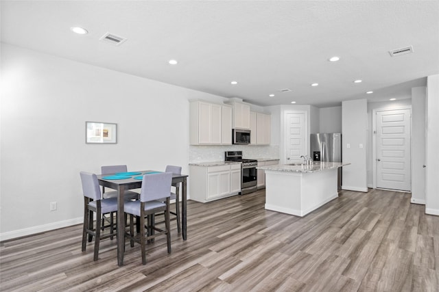 kitchen featuring light wood-type flooring, light stone counters, stainless steel appliances, a kitchen island with sink, and white cabinets