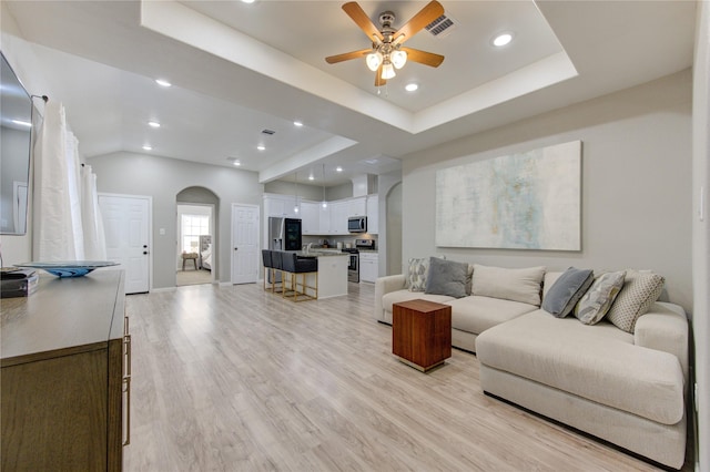 living room with a raised ceiling, ceiling fan, and light wood-type flooring