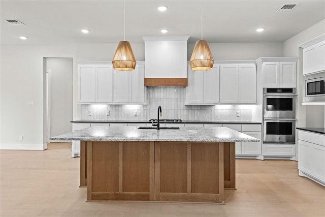 kitchen with light stone counters, premium range hood, decorative light fixtures, a center island with sink, and white cabinets