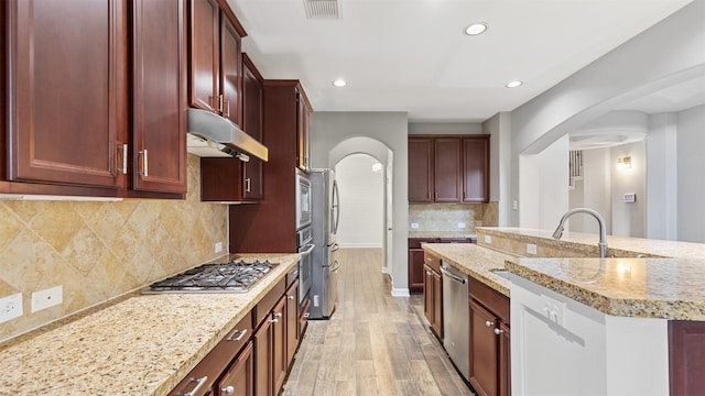 kitchen with stainless steel appliances, tasteful backsplash, light stone counters, light hardwood / wood-style floors, and a center island with sink