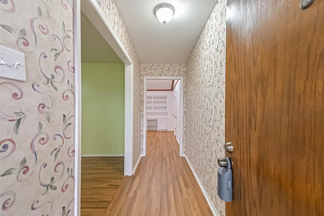 corridor with light wood-style floors, a textured ceiling, baseboards, and wallpapered walls