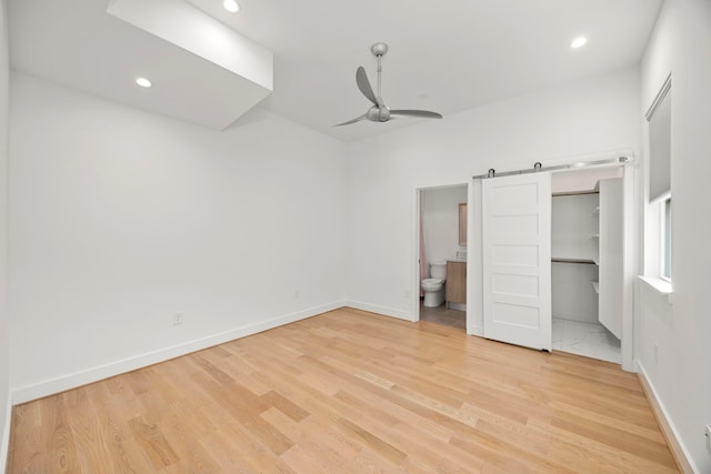 unfurnished bedroom with light hardwood / wood-style floors, connected bathroom, a barn door, and ceiling fan