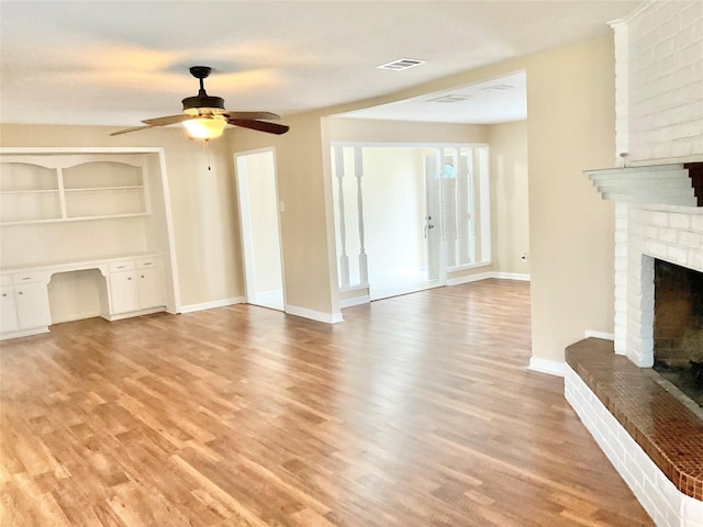 unfurnished living room featuring a brick fireplace, built in desk, light hardwood / wood-style flooring, and ceiling fan