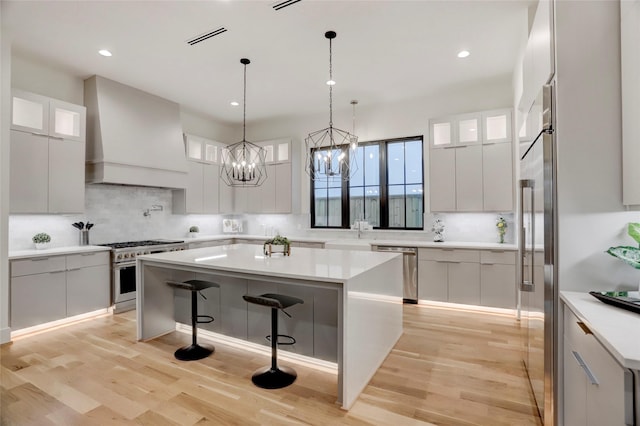 kitchen featuring premium appliances, light hardwood / wood-style floors, premium range hood, and a kitchen island with sink