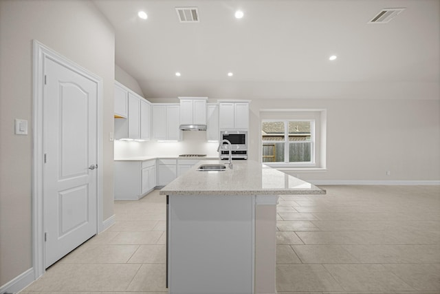 kitchen featuring stainless steel microwave, light stone counters, an island with sink, and white cabinetry