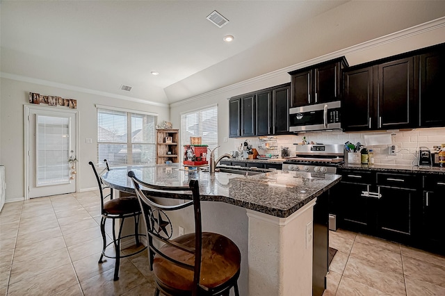 kitchen featuring dark stone counters, a center island with sink, vaulted ceiling, ornamental molding, and appliances with stainless steel finishes