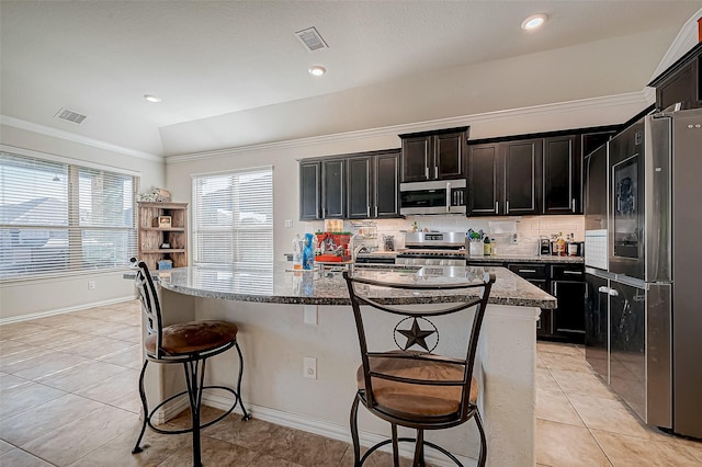 kitchen with light stone counters, an island with sink, vaulted ceiling, light tile patterned floors, and appliances with stainless steel finishes