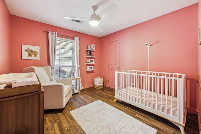 bedroom with a textured ceiling, ceiling fan, a crib, and dark wood-type flooring