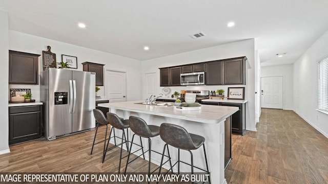 kitchen with hardwood / wood-style floors, a kitchen island with sink, sink, appliances with stainless steel finishes, and dark brown cabinetry