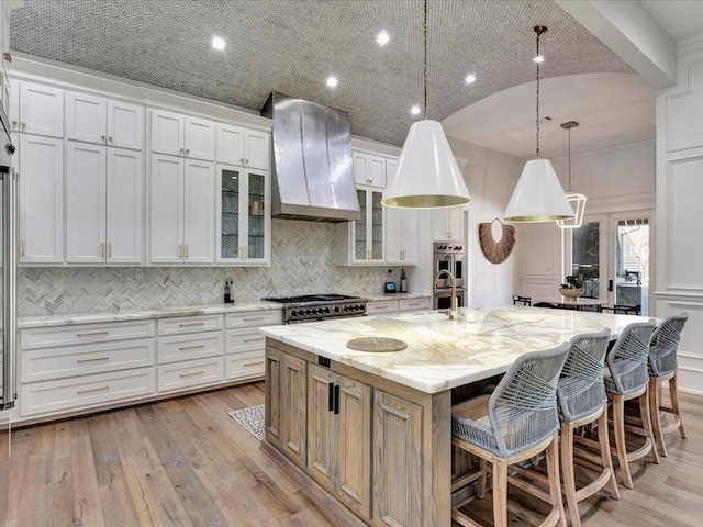 kitchen featuring a large island with sink, wall chimney range hood, light wood-type flooring, light stone counters, and white cabinetry