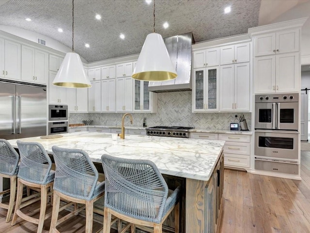 kitchen featuring light stone counters, wall chimney range hood, white cabinets, hanging light fixtures, and an island with sink